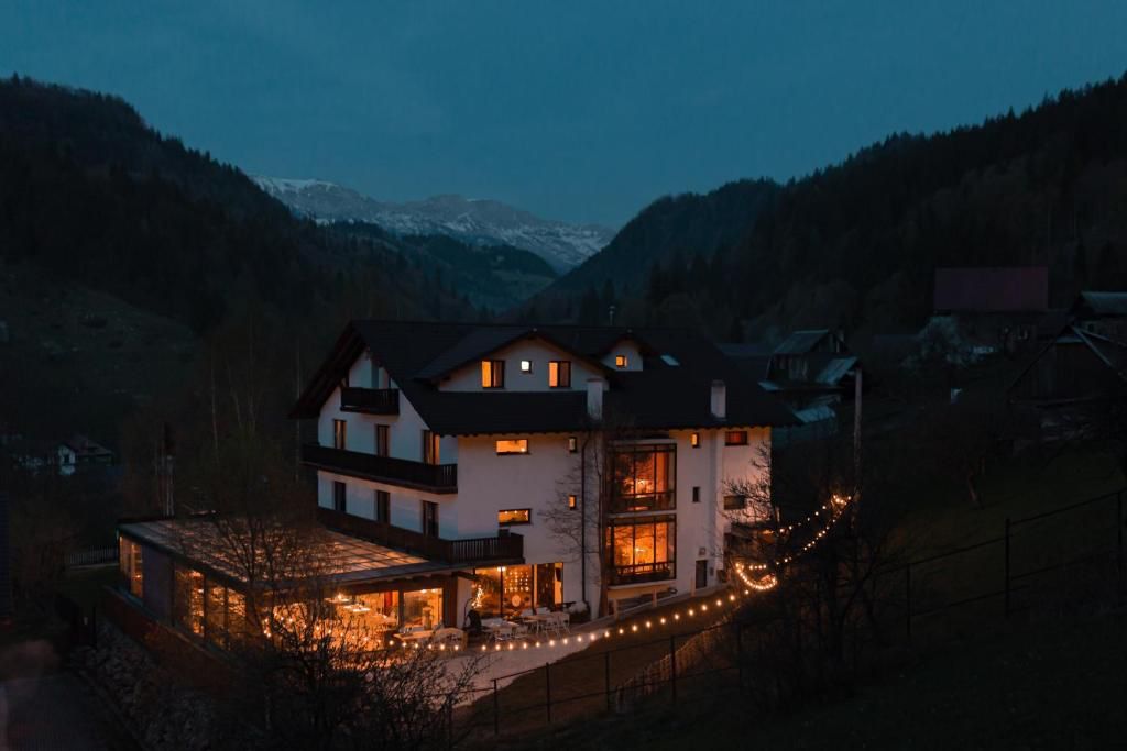 Casa de retreat seara, cu ferestrele calde și șiraguri de lumini pe terasă, între dealuri împădurite