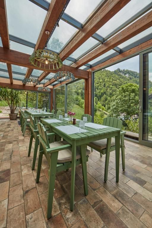 Veranda de sticlă cu masa lungă și scaune verzi, deschisă spre pădure